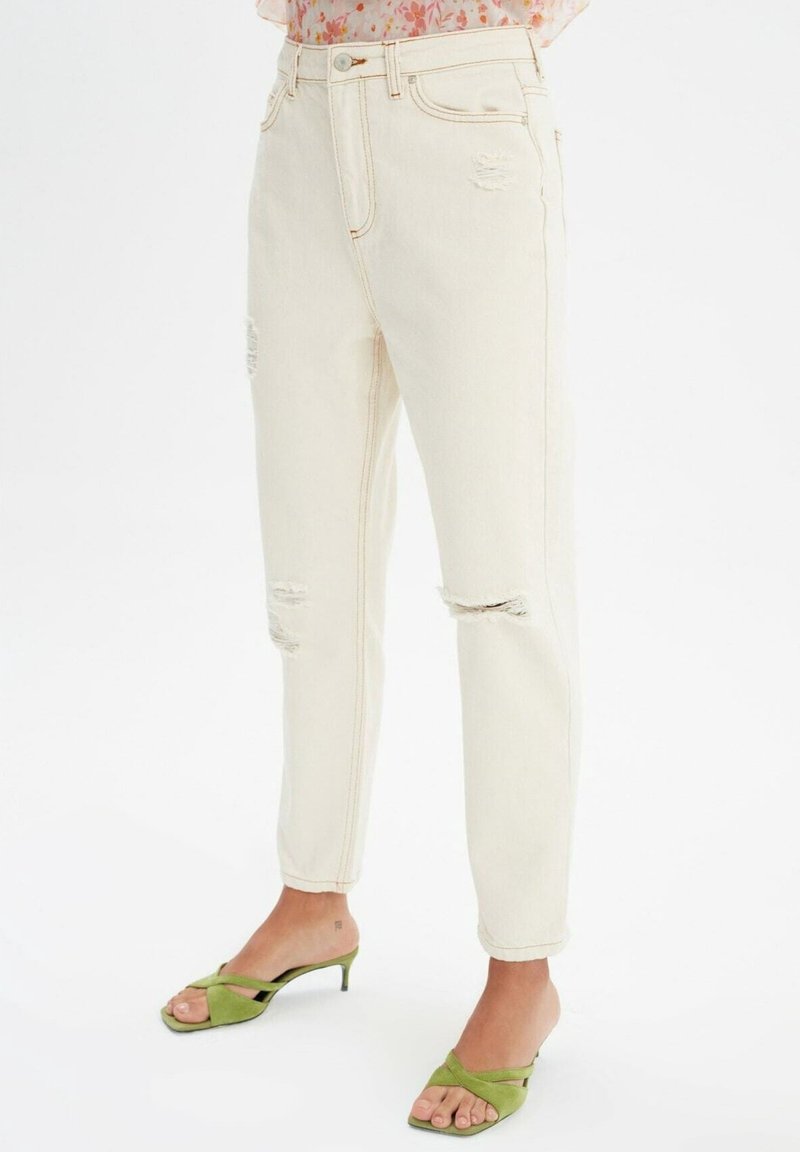 Jeans de tiro alto en beige claro con detalles desgastados y costuras visibles, combinados con tacones abiertos en verde claro.