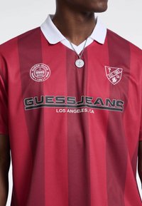 Rot und kastanienbraun gestreiftes Polohemd mit einem weißen Kragen, das das Logo und Embleme "GUESS JEANS" auf der Brust trägt. Aus strukturiertem Stoff gefertigt.