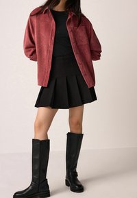 Czerwona koszula sztruksowa na guziki noszona na dopasowanym czarnym topie, zestawiona z czarną plisowaną spódnicą i czarnymi skórzanymi kozakami za kolano.