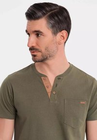 Camisa de manga corta verde oliva con escote tipo henley, acentos de botones marrones y un bolsillo en el pecho. Textura suave y corte relajado.