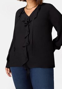 Svart blus med V-ringning, framsidan har volanger och knytning. Långa ärmar med knäppning vid handleden. Bärs med blå denimjeans. Slät textur.