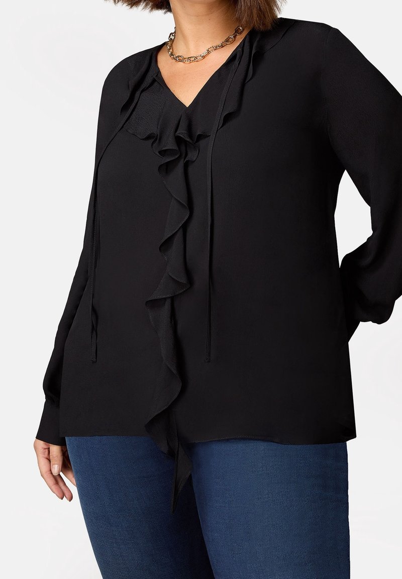 Svart blus med V-ringning, framsidan har volanger och knytning. Långa ärmar med knäppning vid handleden. Bärs med blå denimjeans. Slät textur.