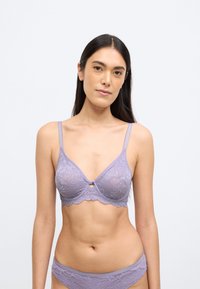 Violetinė nėriniuota braletė su banguotu apvadu, reguliuojamomis petnešėlėmis ir raktikaulio detalė centre. Lygi audinio tekstūra.