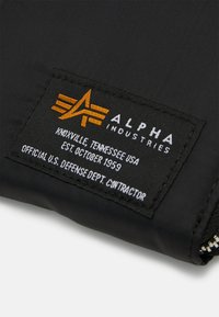 Étiquette en tissu noir avec texte orange et blanc, présente le logo "ALPHA INDUSTRIES", les détails de localisation et "CONTRACTANT OFFICIEL DU DEPARTEMENT DE LA DEFENSE DES ÉTATS-UNIS."