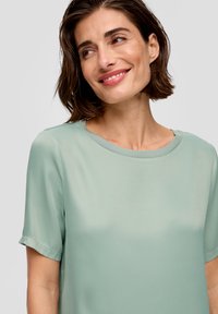 Hellgrüne Bluse aus glattem Stoff, mit kurzen Ärmeln, Rundhalsausschnitt und lockerer Passform. Minimalistisches Design ohne Muster.