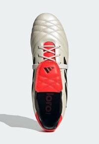 Zapato de fútbol gris claro con parte superior de cuero texturizado, acentos rojos, franjas laterales negras y cordones visibles, así como una marca en la plantilla.