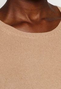 Beiger Strickpullover mit rundem Ausschnitt und ribbeltem Rand. Die Textur ist weich und glatt, was das feine Strickmuster hervorhebt.