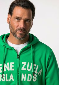 Sudadera verde con cremallera y la inscripción "ZUELA ISLANDS" en blanco. Cuenta con capucha, cordones y una textura suave, ideal para el uso casual.