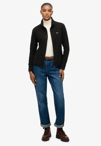 Zwarte fleecejas met hoge kraag, crèmekleurige coltrui, blauw opgerolde jeans en bruine veterlaarzen. Model staat recht naar voren gericht.