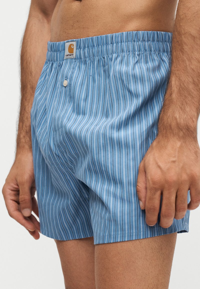 Mann trägt blau-weiße, vertikal gestreifte Boxershorts mit elastischem Bund und vorderem Knopfverschluss, gezeigt von der Taille bis zur Mitte des Oberschenkels.