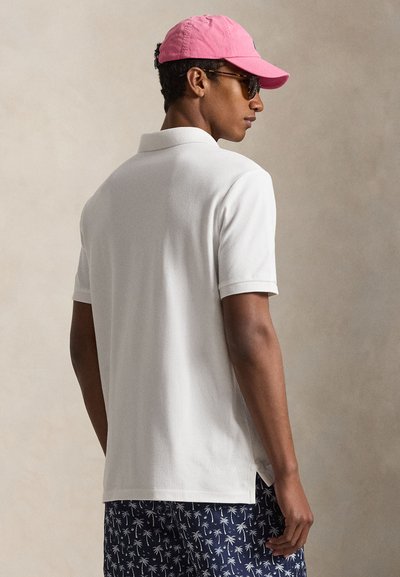 Mann iført hvit polo skjorte, marineblå shorts med hvitt palmemønster, rosa baseballcaps og brune solbriller, sett fra siden mot en ensfarget bakgrunn.