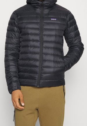Mann trägt eine schwarze gesteppte Daunenjacke von Patagonia mit Kapuze und beige Hose, eine Hand in der Tasche, vor neutralem Hintergrund.