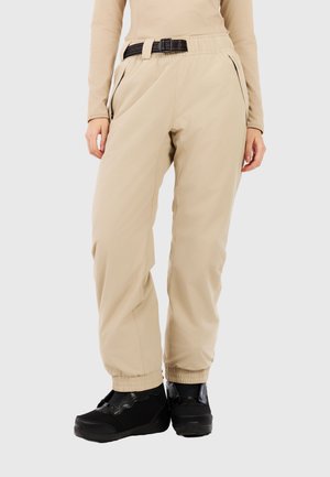 Person iført beige elastiske bukser med sideslidslommer og sort bælte, sat sammen med sorte atletik sko og beige langærmet top.