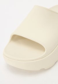 Ljusbeige slide-sandal med öppen tå, smidigt gummimaterial, texturerad fotbädd och något upphöjd plattformsula.