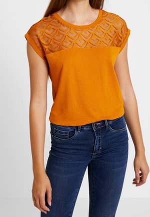Kvinde iført orange kortærmet top med geometrisk blonderyg og blå denimjeans, stående foran en ensfarvet baggrund.