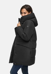 Woman wearing a black oversized puffer jacket with high collar and hood, posing sideways against a plain white background.