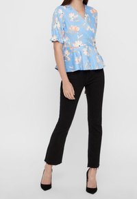 Top peplum azzurro chiaro con motivo floreale e maniche corte a volant, abbinato a pantaloni neri aderenti e tacchi alti neri.