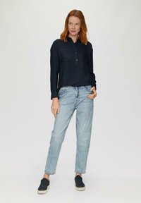 Marineblaues Button-Up-Hemd mit zwei Brusttaschen, kombiniert mit hellblauen, geraden Jeans und dunklen Sneakers, stehend vor einem neutralen Hintergrund.