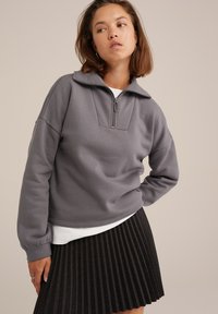 Grå kvart lynlås sweatshirts med høj krave og tekstureret panel; båret over en hvid skjorte. Kombineret med en sort plisseret nederdel.