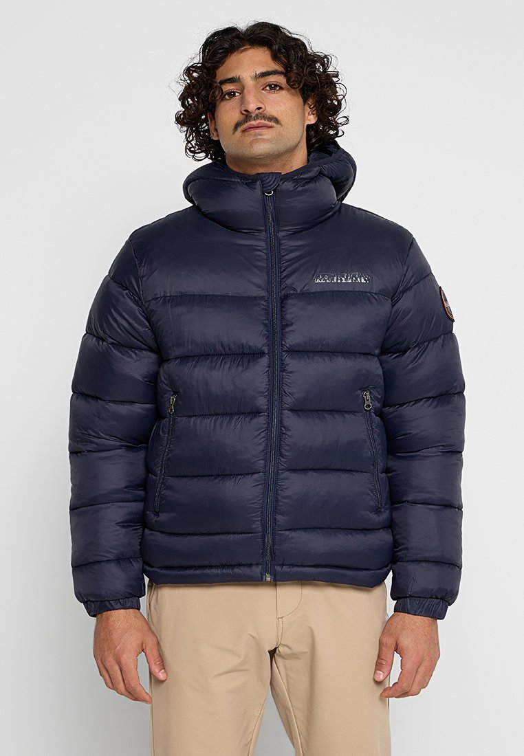 Mann mit lockigem Haar und Schnurrbart, der eine navyblaue Steppjacke und beigefarbene Hose trägt, steht vor einem schlichten, hellen Hintergrund.