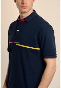 Polo de color marino hecho de algodón con cuello extendido, que presenta rayas horizontales rojas y amarillas, y un logo en el pecho.