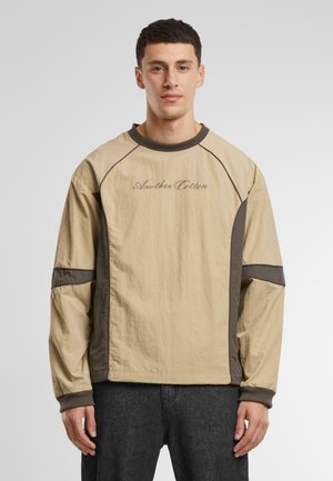 Beige long-sleeve top with dark gray accents, round neck, and ‘Another Cotton’ logo. Textured fabric with a relaxed fit and raglan sleeves.