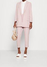 Lichtroze blazer met bijpassende cropped broek, gecombineerd met een wit blouse en witte platte sandalen. Draagt een tan geknoopte tas.