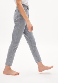Jeans in denim grigio con vita aderente, gambe affusolate e orlo grezzo. Il tessuto ha una finitura strutturata e chi li indossa è a piedi nudi.