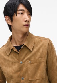 Rjava semiš jakna s ošiljenim ovratnikom, srebrnimi gumbi na pritrditev in žepom na prsih s majhnim logotipom. Mehak material, krojen fit.