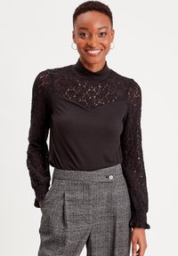 Haut noir à manches longues avec des détails en dentelle sur les manches et un col montant, associé à un pantalon à carreaux gris avec des plis et un bouton.