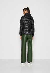 Giacca nera trapuntata con cappuccio, caratterizzata da un motivo a onde e polsini elastici, abbinata a pantaloni in pelle verde e sneakers nere.