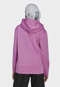 Sweatshirt à capuche rose avec une texture lisse. Il dispose d'une poche kangourou et de poignets côtelés, stylé avec un foulard blanc.