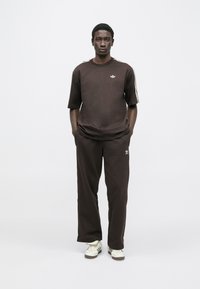 Brown cotton t-shirt with short sleeves and white logo, paired with matching brown trousers. The outfit includes white sneakers with black accents.