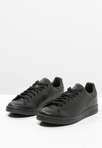 Černé kožené tenisky s hladkou texturou, zaoblenou špičkou, perforacemi pro prodyšnost a černou gumovou podrážkou. Mají rovné tkaničky.