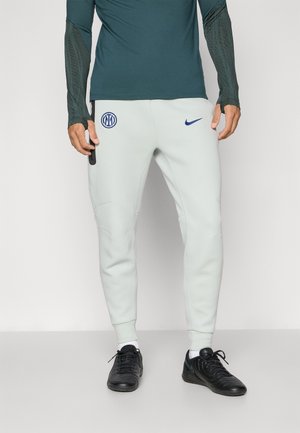 Lysegrå Nike sweatpants har en tætsiddende pasform, sidelommer og et blåt logo på venstre lår, parret med sorte træningssko.