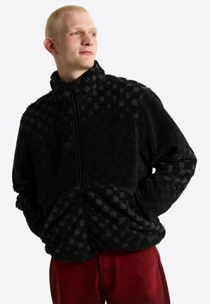Junge Person mit kurzen blonden Haaren, die eine schwarze, strukturierte Reißverschlussjacke und rote Hose trägt, schaut zur Seite mit den Händen in den Taschen.