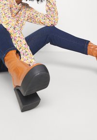 Bruine kniehoge laarzen met hak, vierkante neus en textuursula, gecombineerd met donkerblauwe spijkerbroek en een meerkleurige top met bloemmotief.