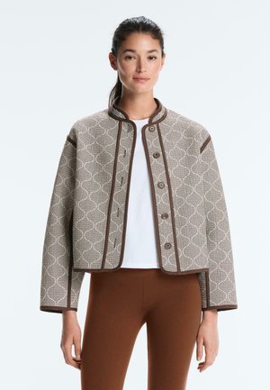 Femme portant une veste beige à motifs boutonnée avec des bordures marron sur un haut blanc et un pantalon marron ajusté, debout devant un fond uni.