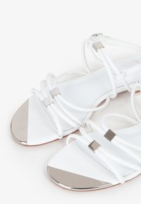 Sandali bianchi con cinturini, upper in pelle liscia, dettagli metallici sulla punta e tacco piatto. Presentano un design minimalista e punta aperta.