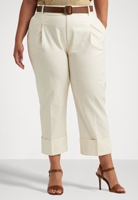 Nainen, joka on pukeutunut vaalean kermaisiin lyhyisiin housuihin, joissa on käärityt lahkeet, ruskeaan vyöhön ja ruskeisiin korkeakorkoisiin sandaaleihin, seisoo kädet taskuissa.