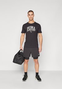Camiseta deportiva negra con "PUMA sports club" impreso, combinada con pantalones cortos negros, zapatillas negras y sosteniendo un bolso de deporte negro.