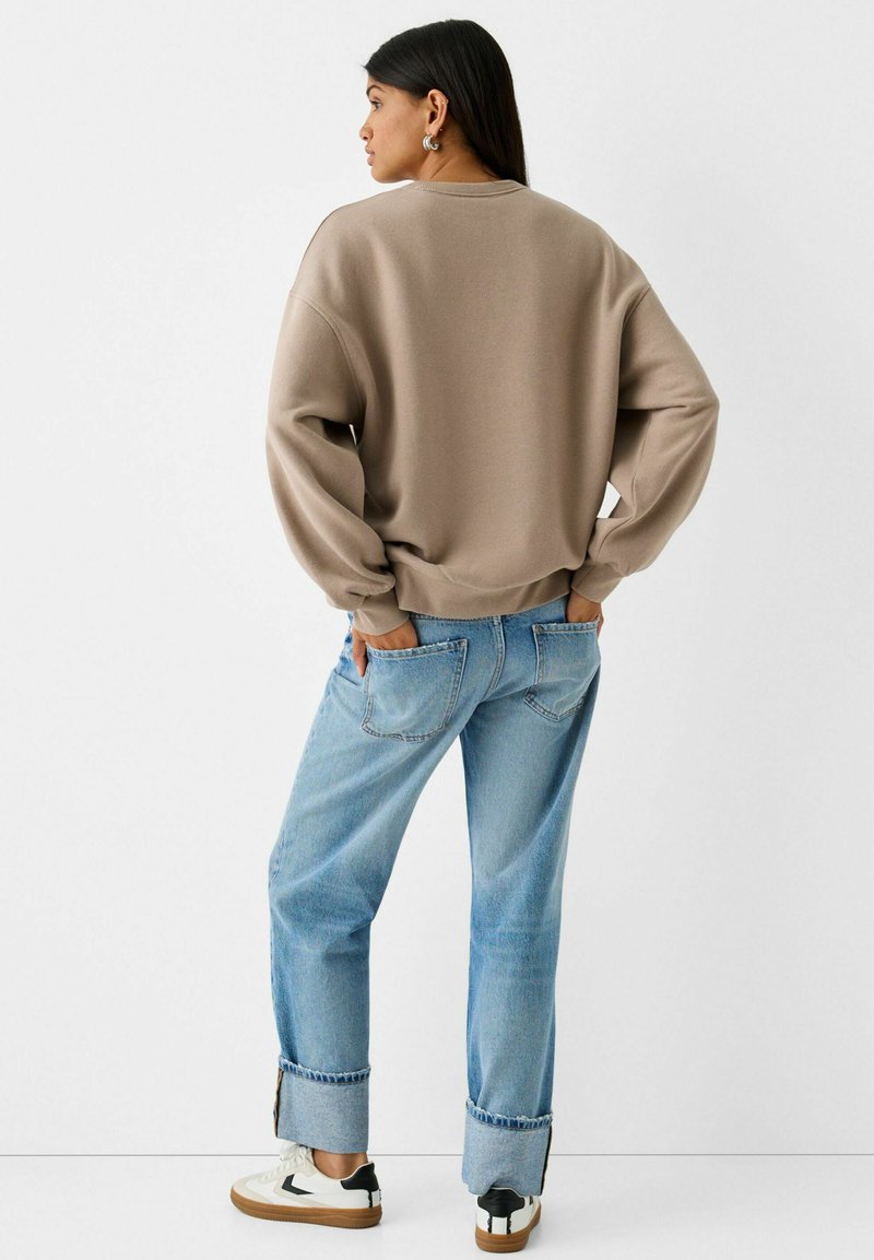 Donna con le mani nelle tasche posteriori, che indossa una felpa beige, jeans azzurri arrotolati, sneakers bianche e orecchini d'argento, rivolta di lato.