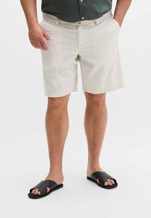 Mann trägt beige Shorts, dunkelgrünes Hemd und schwarze Slide-Sandalen, steht mit den Händen in den Taschen vor weißem Hintergrund.