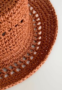 Sombrero de sol tejido de terracota con ala ancha, que presenta un patrón de crochet texturizado y pequeños detalles recortados a lo largo del borde.