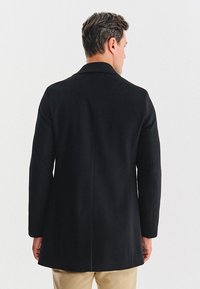 Czarny wełniany płaszcz z pojedynczym tylnym rozcięciem, dopasowany krój, klapy w kształcie litery V i gładka faktura. Minimalistyczny design z wyraźnymi liniami.