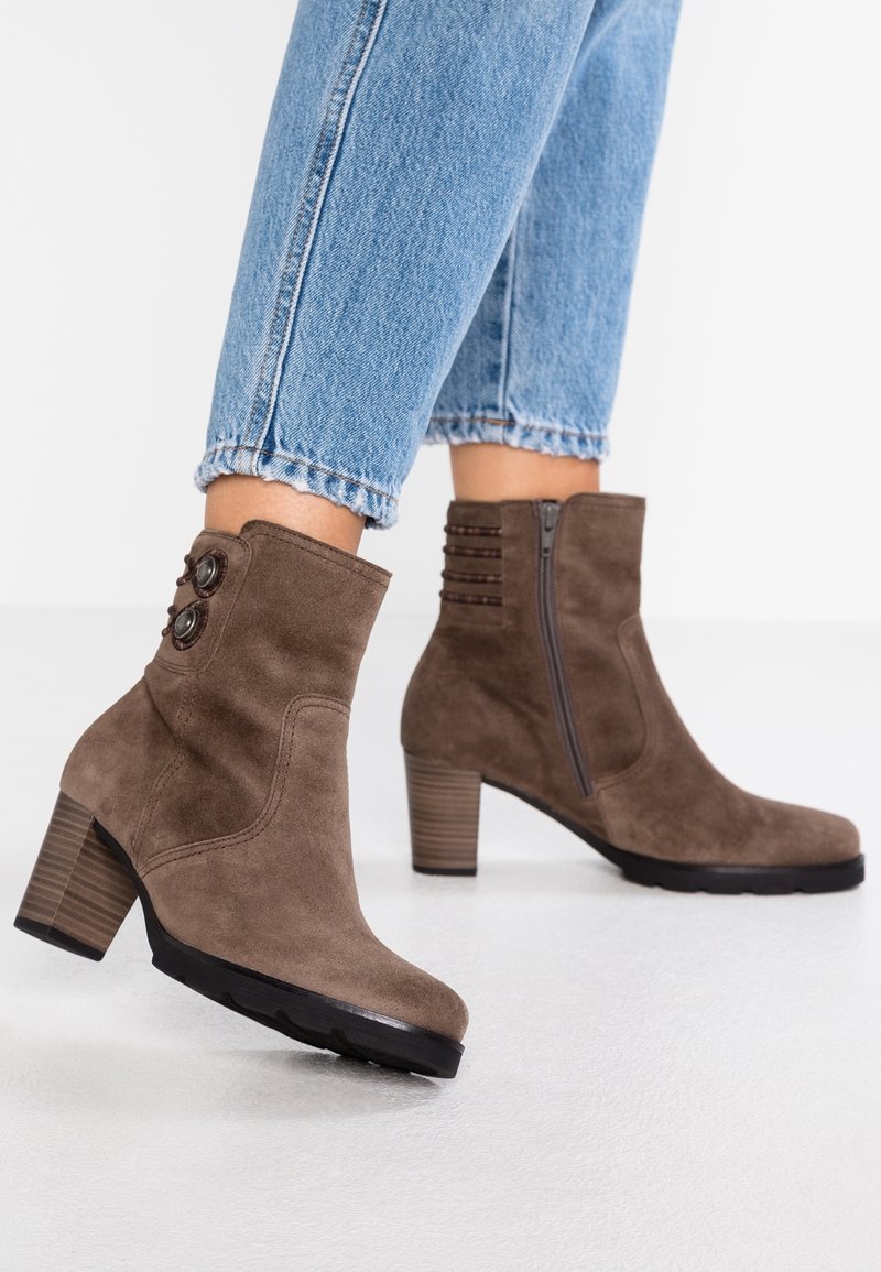 Bottes basses en daim marron avec talons empilés et fermeture éclair latérale, portées avec un jean bleu retroussé sur fond blanc.