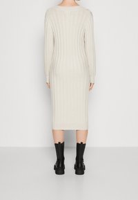 Vrouw gezien vanuit de rug, gekleed in een lange crèmekleurige geribbelde gebreide jurk en zwarte chunky laarzen tot halverwege de kuit, staande tegen een egale witte achtergrond.