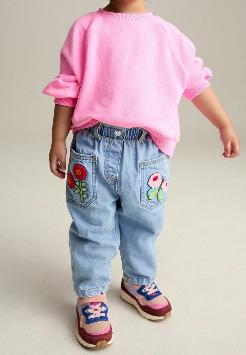 Tout-petit portant un haut à manches longues rose, un jean bleu clair avec des patchs de fleurs et de papillons sur les poches, et des baskets roses et bleues.