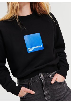 Kobieta w czarnej bluzie z niebieskim kwadratem i napisem „KARL LAGERFELD JEANS”, w ciemnoszarych dżinsach, ręce w kieszeniach.