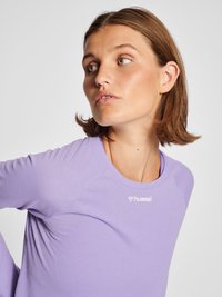 Vrouw met kort bruin haar die een lichtpaars hummel sportshirt draagt, kijkt naar links tegen een effen witte achtergrond.